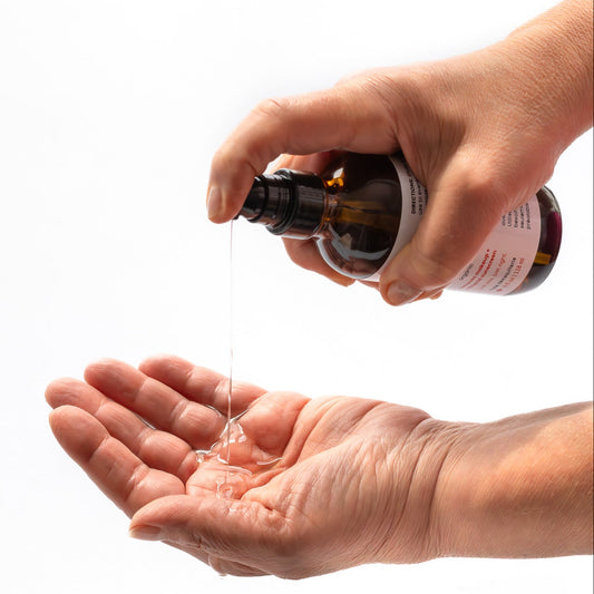 Hand holding amber 4 oz glass pump bottle of 'the cleansing oil', pouring the oil into another hand on a white background
