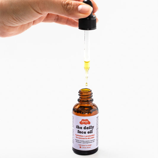 Hand holding a dropper over a bottle of 'the daily face oil' with a white background